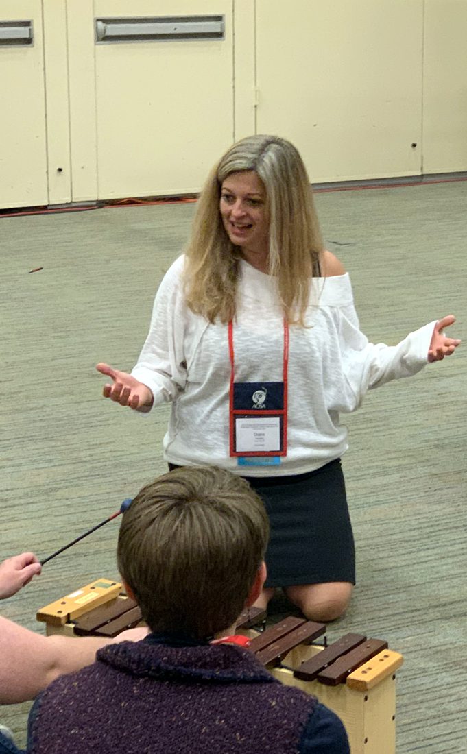 Diana Hawley kneeling in a circle of music teachers, arms open, a xylophone in front of her